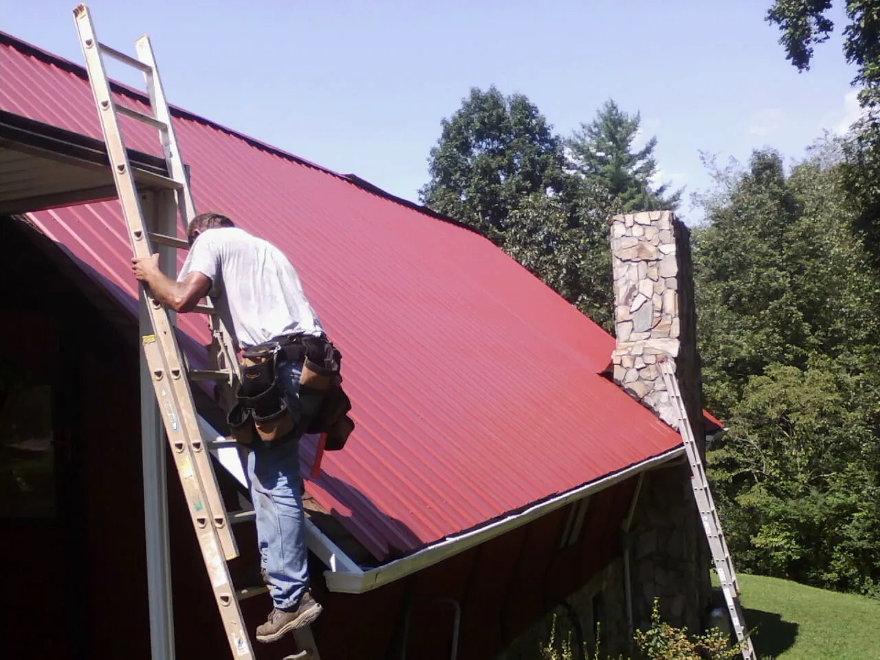 Roofer on a ladder beside a red metal roof and stone chimney — Soffit & Fascia Repair by Roofing Brothers in Front Royal